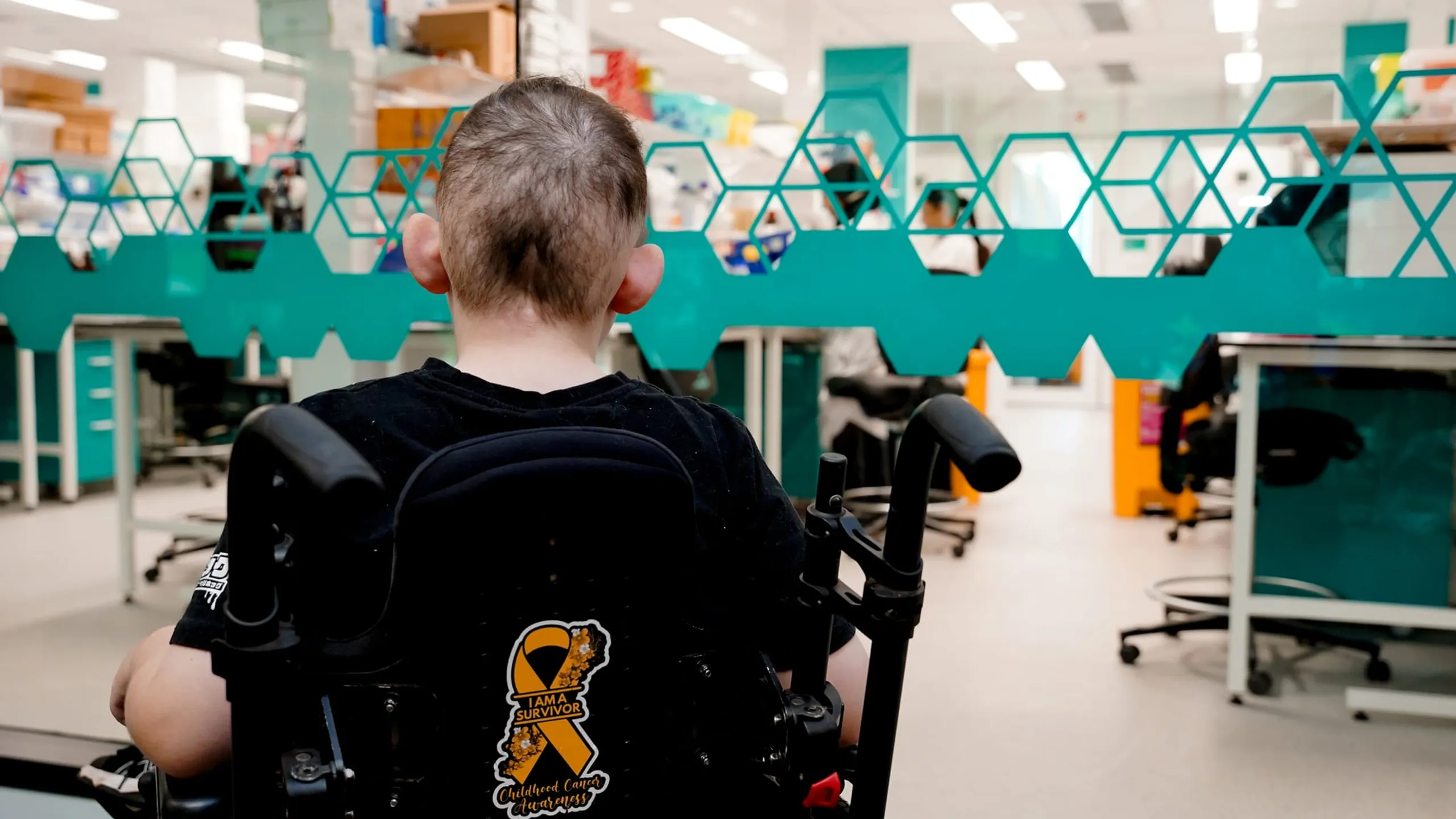 A child in a wheelchair looks through a glass wall into a medical research laboratory.