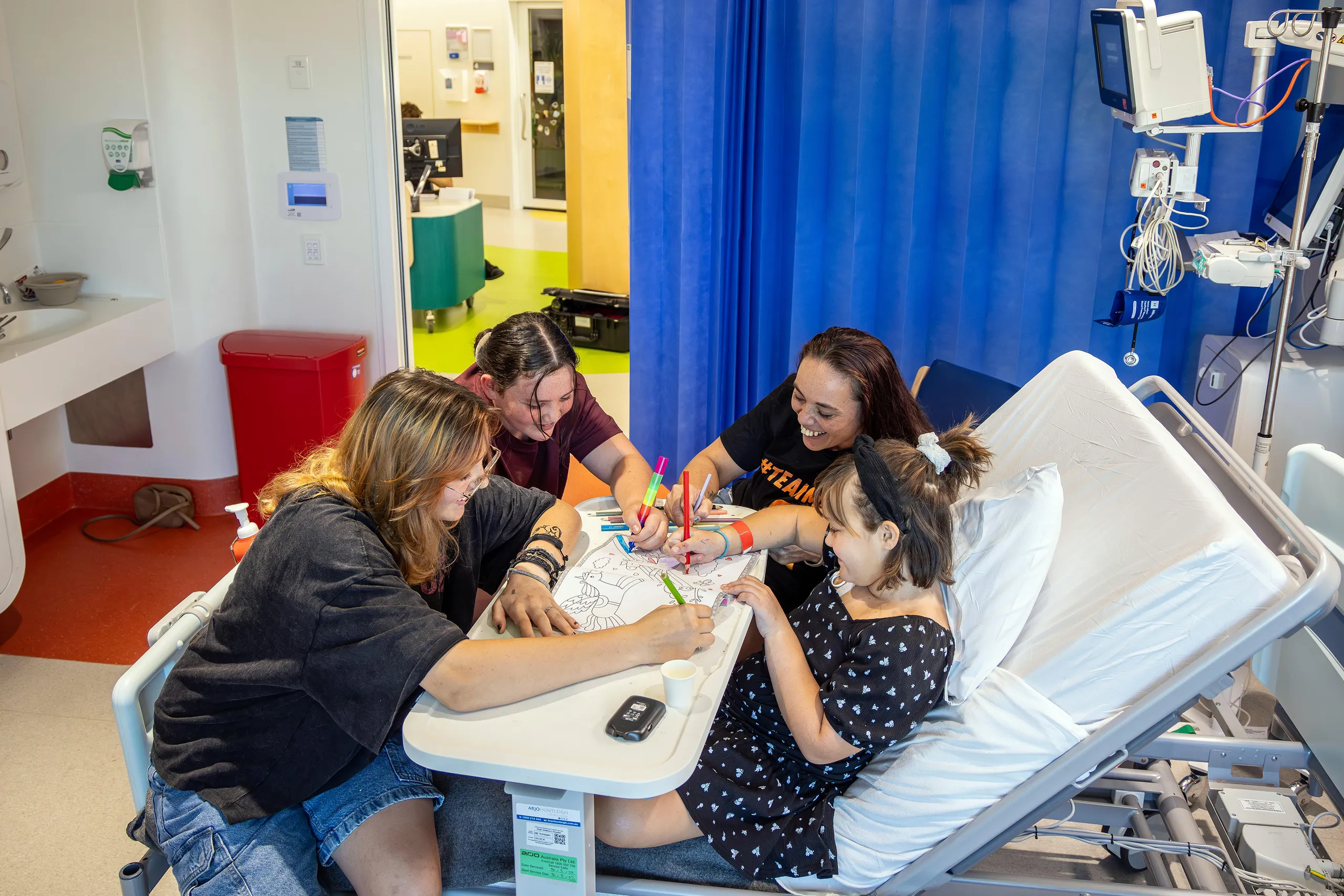 A young hospital patient sits in a hospital bed colouring with family members around a bedside table in a paediatric ward room.