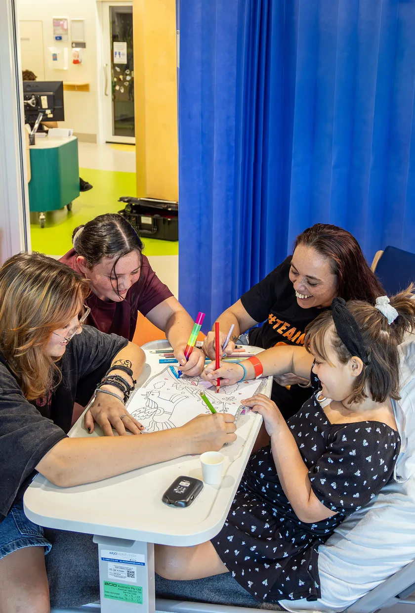 A young hospital patient sits in a hospital bed colouring with family members around a bedside table in a paediatric ward room.