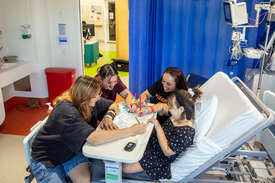 A young hospital patient sits in a hospital bed colouring with family members around a bedside table in a paediatric ward room.