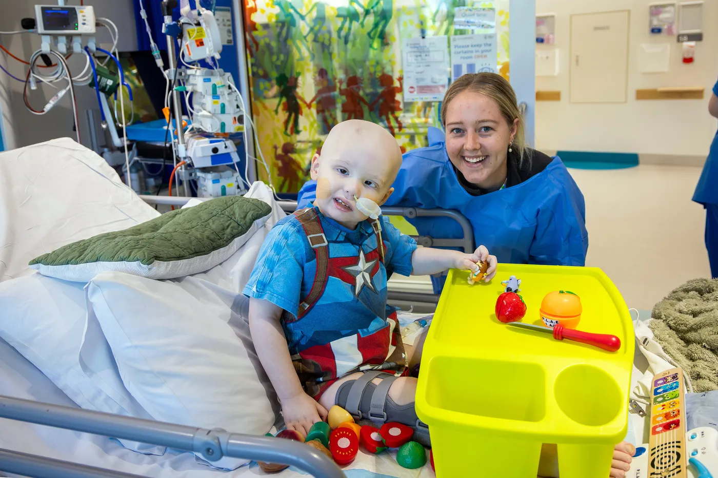 A young hospital patient sits in a hospital bed playing with toys while a healthcare worker smiles beside them in a paediatric ward room.