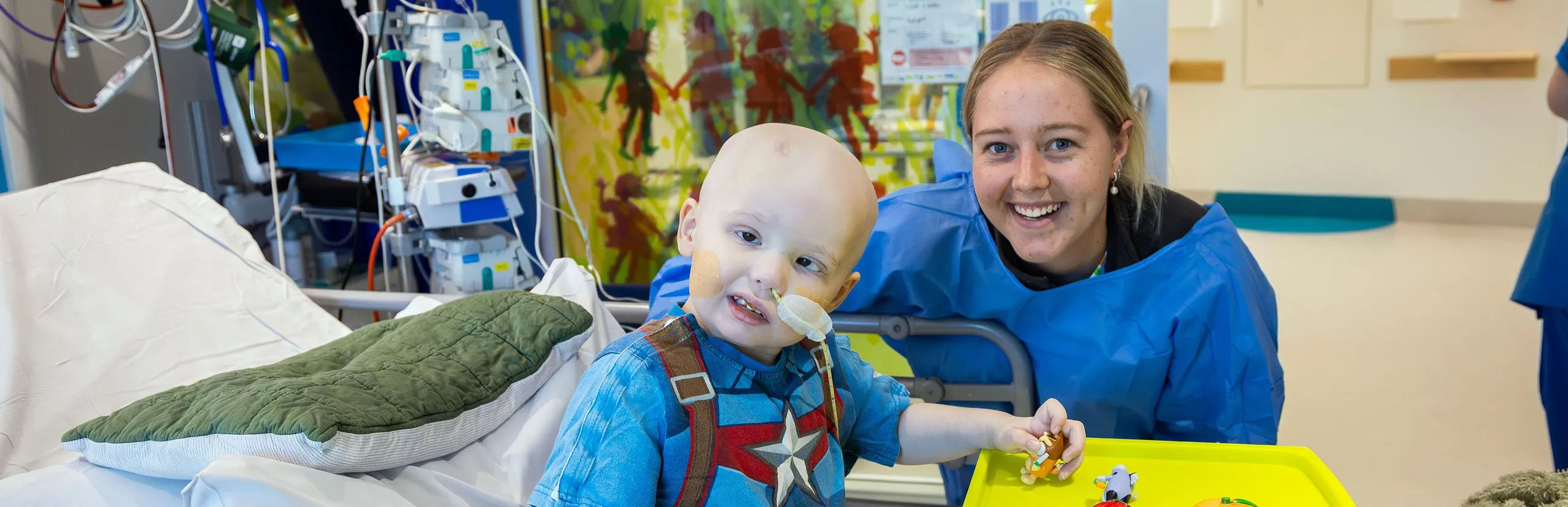 A young hospital patient sits in a hospital bed playing with toys while a healthcare worker smiles beside them in a paediatric ward room.