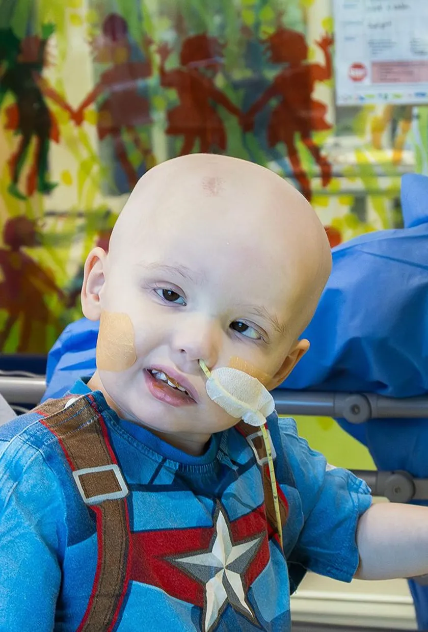 A young hospital patient sits in a hospital bed playing with toys while a healthcare worker smiles beside them in a paediatric ward room.