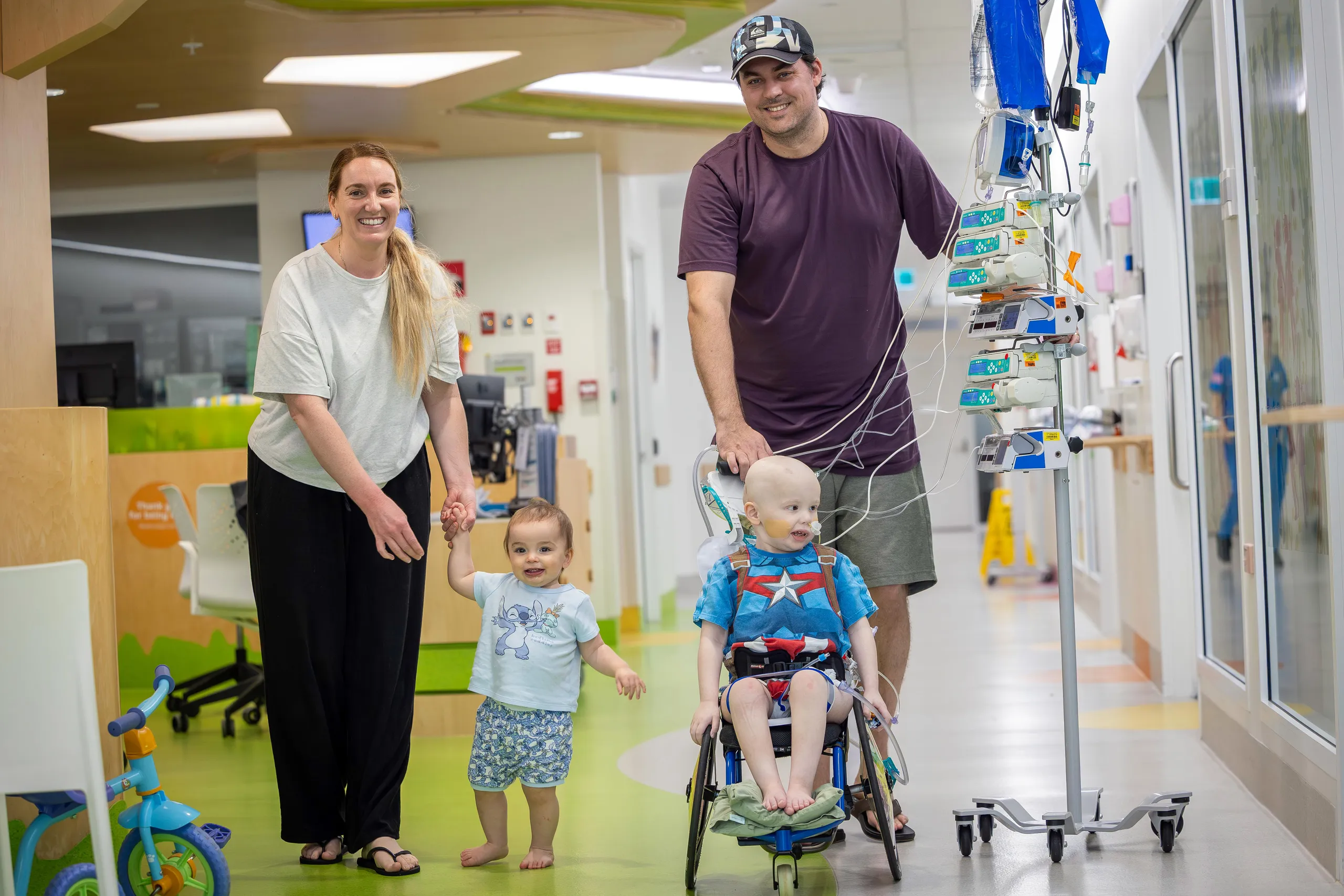A child receiving treatment sits in a wheelchair connected to medical equipment while smiling parents and a younger sibling walk together through a hospital corridor.