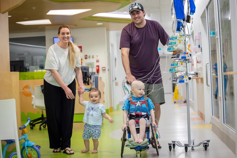 A child receiving treatment sits in a wheelchair connected to medical equipment while smiling parents and a younger sibling walk together through a hospital corridor.