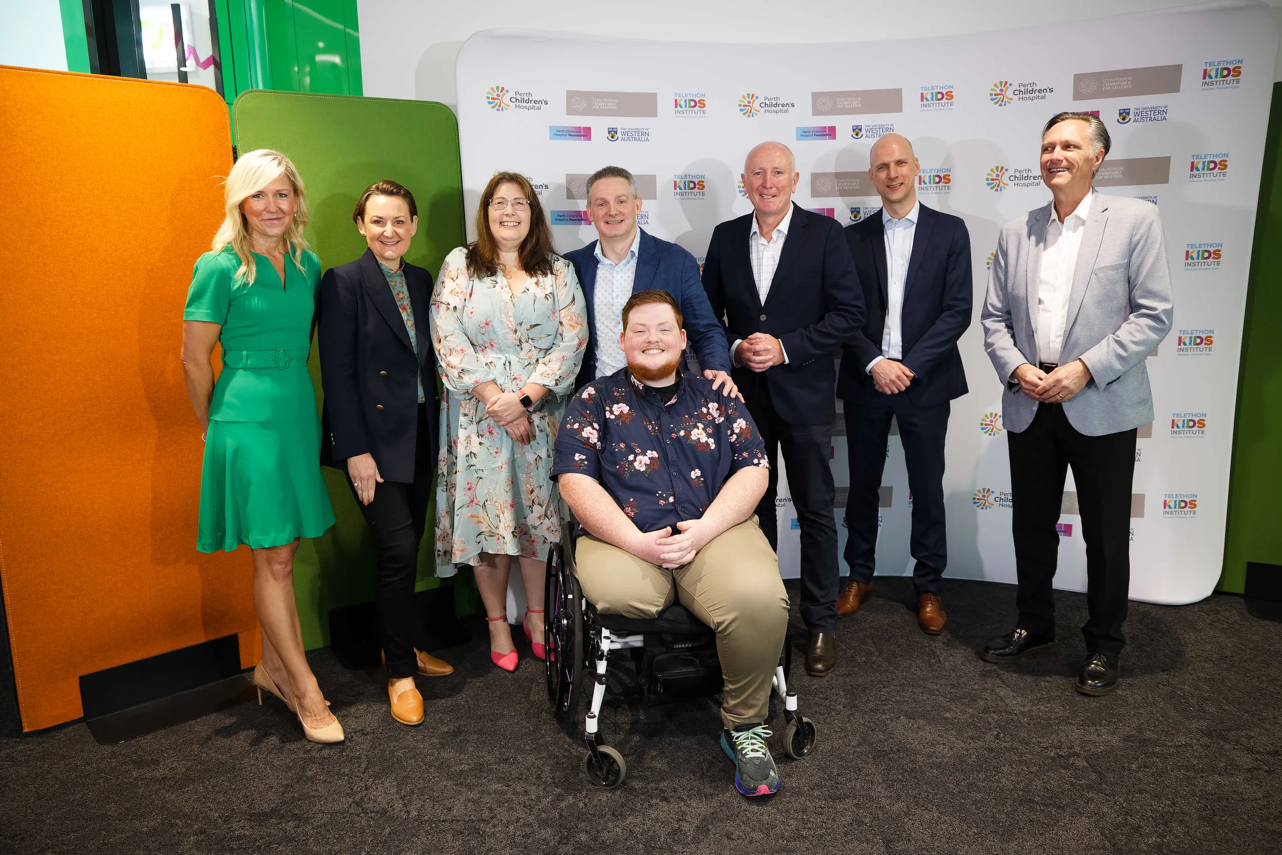 A group of people pose for a photo at a hospital and research institute event, with one attendee seated in a wheelchair in front of a branded media backdrop.
