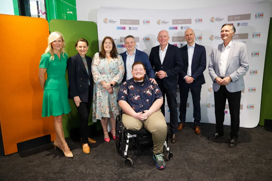 A group of people pose for a photo at a hospital and research institute event, with one attendee seated in a wheelchair in front of a branded media backdrop.