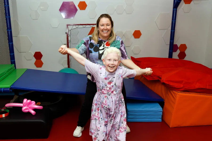 A child and healthcare worker smile and hold hands while playing together in a brightly coloured therapy and activity room.