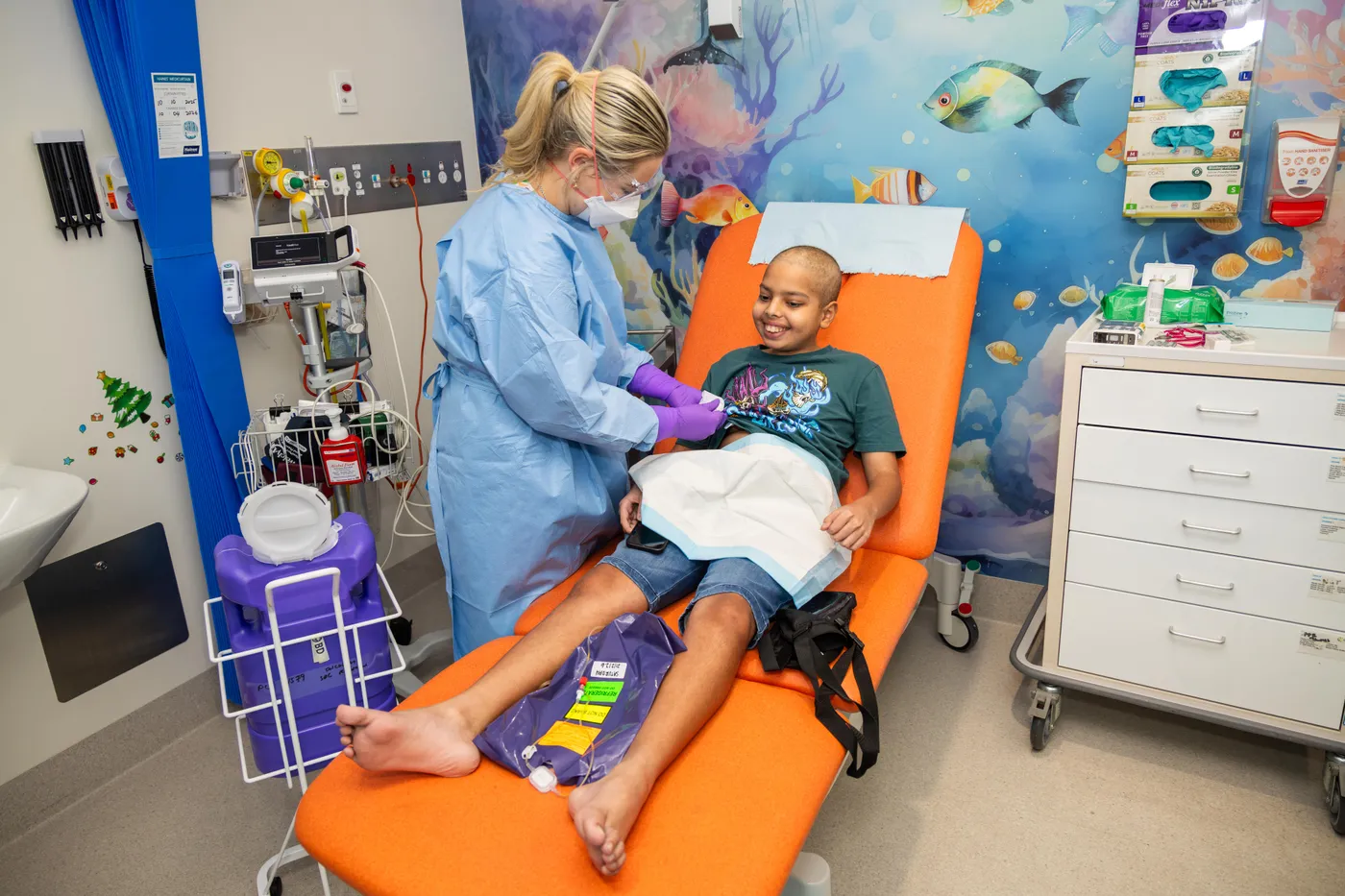 A healthcare worker provides treatment to a smiling young patient seated in a clinic chair in a colourful paediatric treatment room.