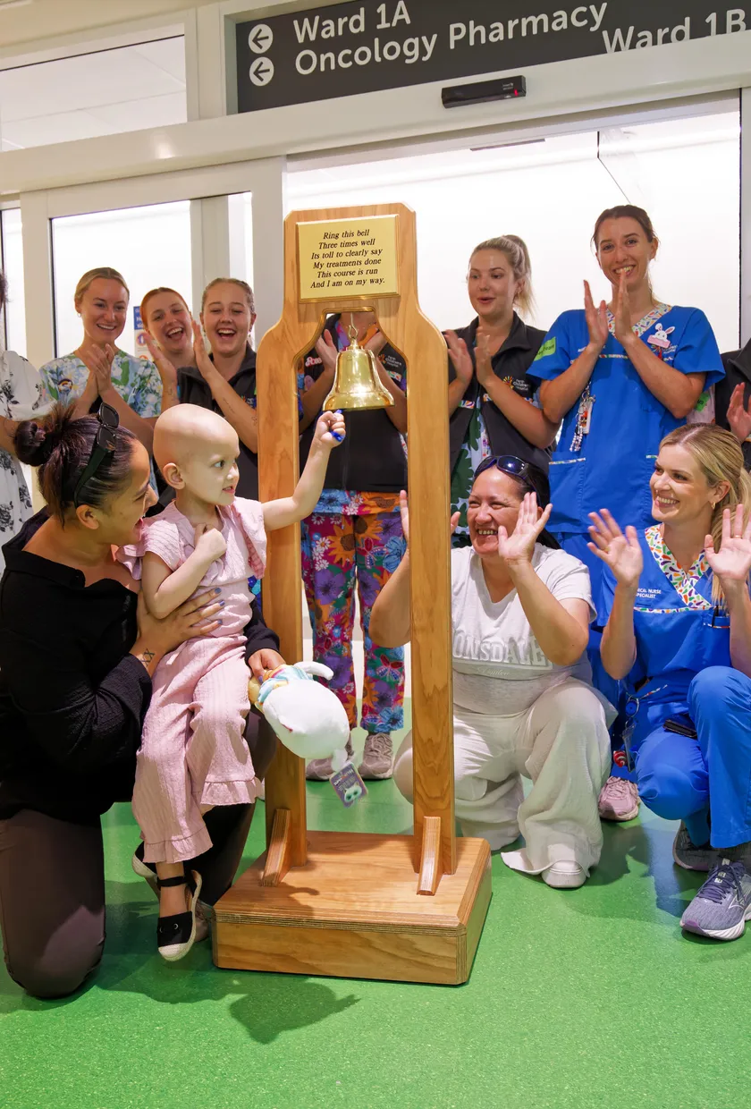 A young child in a hospital corridor rings a ceremonial bell to mark the end of cancer treatment, surrounded by smiling family members, nurses and hospital staff clapping and celebrating.