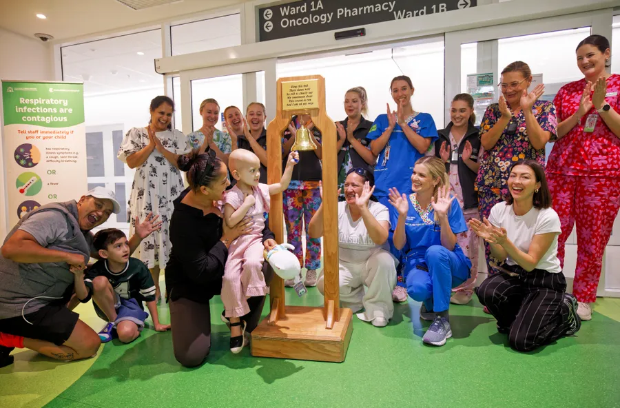 A young child in a hospital corridor rings a ceremonial bell to mark the end of cancer treatment, surrounded by smiling family members, nurses and hospital staff clapping and celebrating.