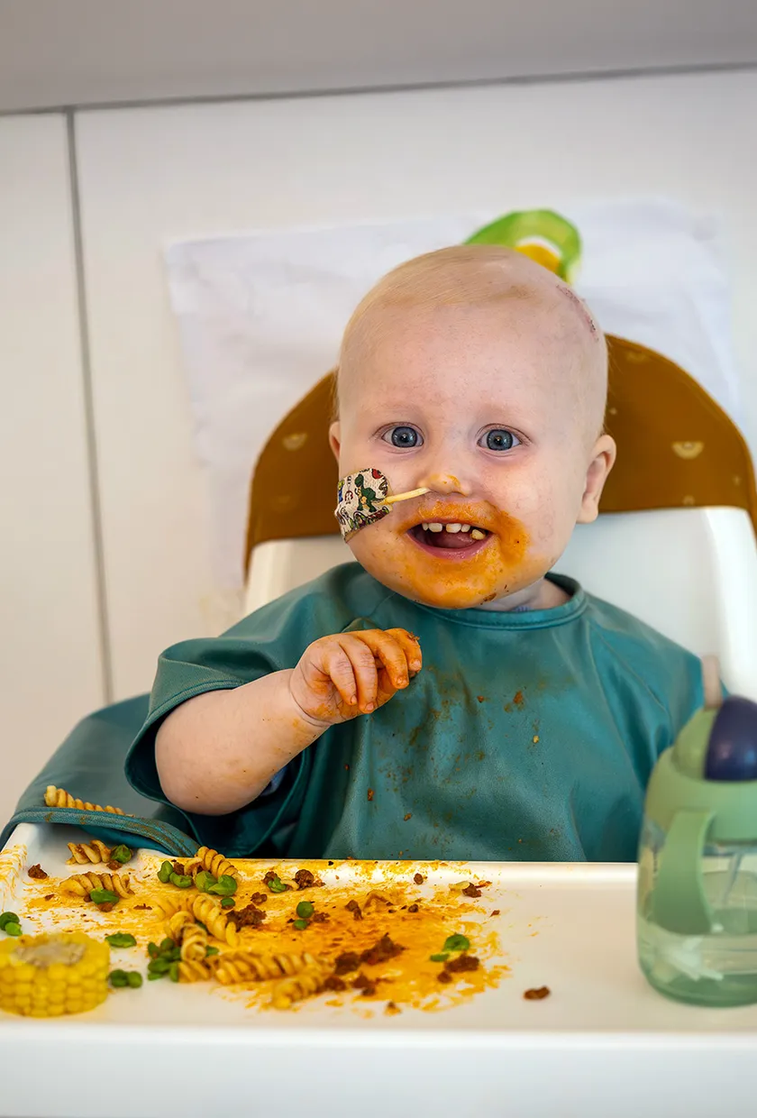 A little baby with a nose tube in a high chair eating food with a big smile.