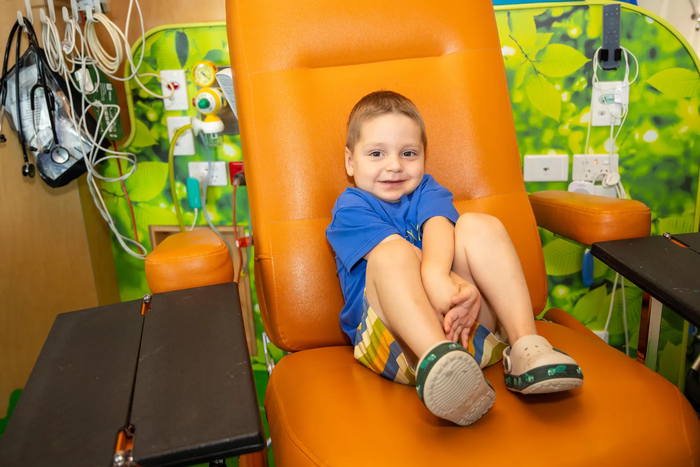 A young child smiles while sitting in a treatment chair in a colourful paediatric hospital room.
