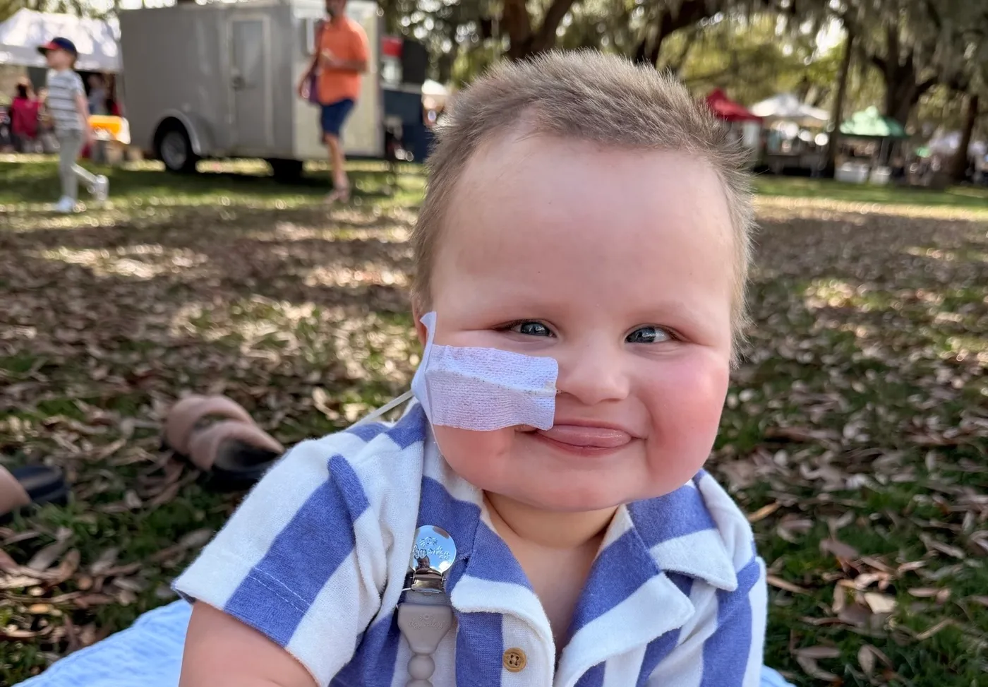 A little boy with a nose tube smiling and sitting in a park.