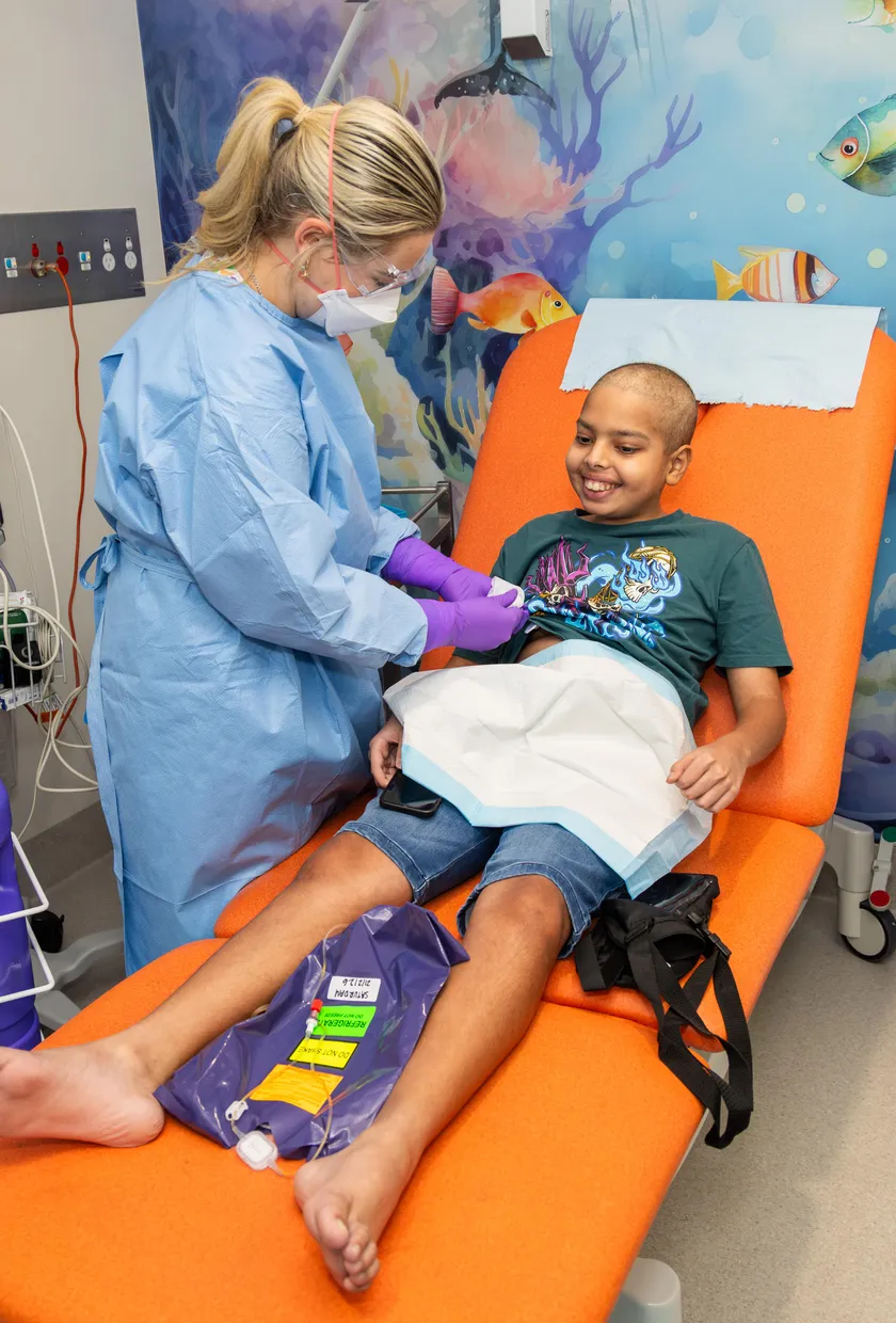 A healthcare worker provides treatment to a smiling young patient seated in a clinic chair in a colourful paediatric treatment room.
